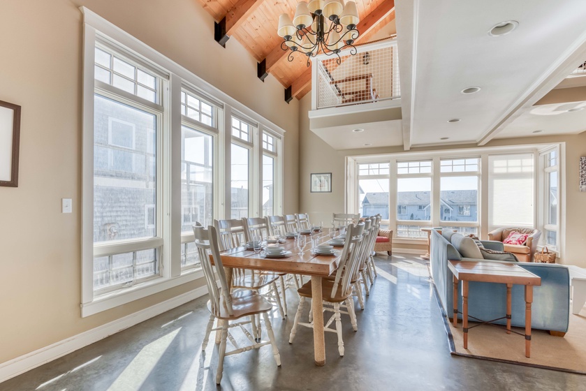 Beach house open living room, Nantucket style, huge dining room table 2 walls of windows, open overhead loft, and a light blue sofa.