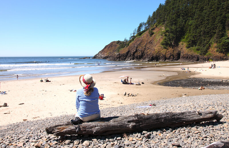 A woman sitting on a log at the beach watching the surfers and people playing in the sand.