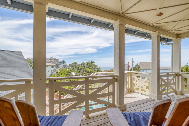 Beach house large covered deck with craftsmen style cream colored railings and 2 Adirondack chairs facing a distant ocean view.