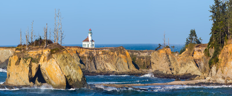 Panoramic view of lighthouse sitting cliffside surrounded by blue sky and ocean.